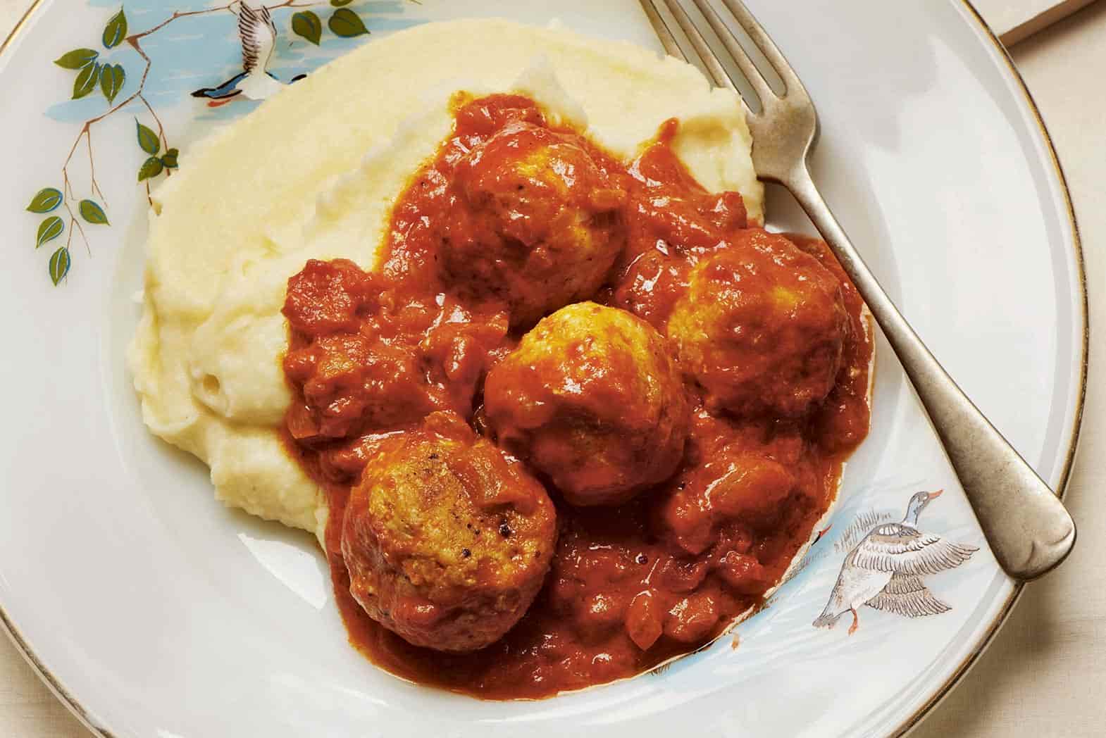 A white plate with bird motif, topped with creamy mashed potato and 4 fish balls in a red tomato sauce